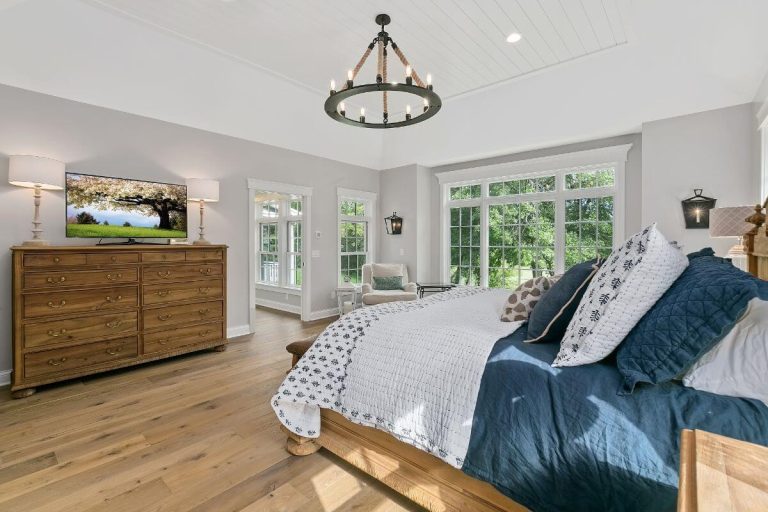 Bright master bedroom with hardwood floors, wooden dresser, bed with blue pillows, and large windows overlooking greenery