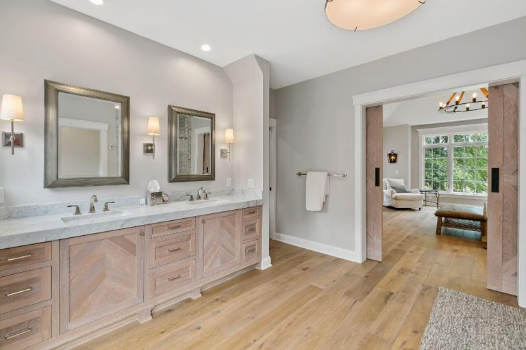 Luxurious master bathroom with dual vanities, large mirrors, wooden cabinetry, and view into bedroom