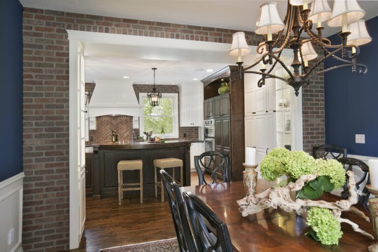 Dining room with exposed brick wall, wooden table, chandelier, and open doorway leading to kitchen area