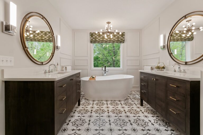 Luxurious bathroom with dual dark vanities, oval mirrors, freestanding tub, and patterned tile flooring