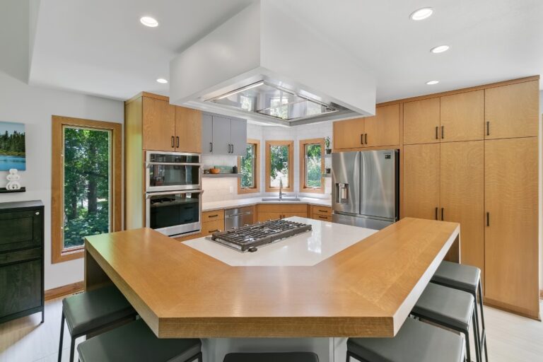 Modern kitchen with wood cabinetry, large island with cooktop, stainless steel appliances, and white ceiling with skylights