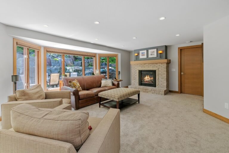 Modern living room with beige carpet, cream furniture, fireplace, large windows, and wood accents