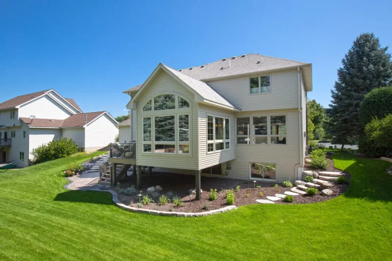 Two-story house with deck and stone patio on manicured lawn surrounded by trees under clear blue sky
