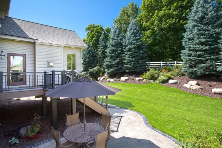 Backyard patio area with deck, seating, lush green lawn, and evergreen trees beside a residential house