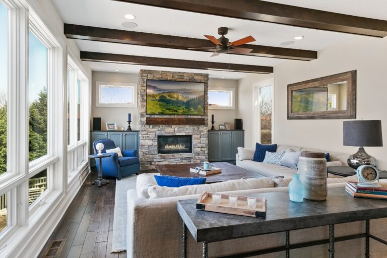 Modern living room with fireplace, blue upholstered furniture, exposed wooden beams, and large windows overlooking outdoor