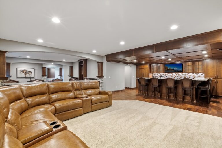 Spacious basement recreation room with tan leather reclining sectional sofa, wood paneling, bar counter with seating, and