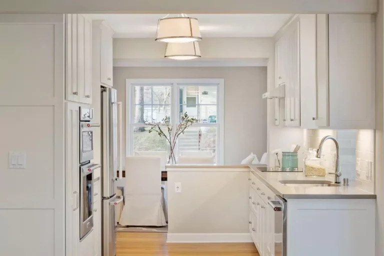 Modern white kitchen with stainless steel appliances, white cabinetry, pendant lighting, and view of backyard through glass