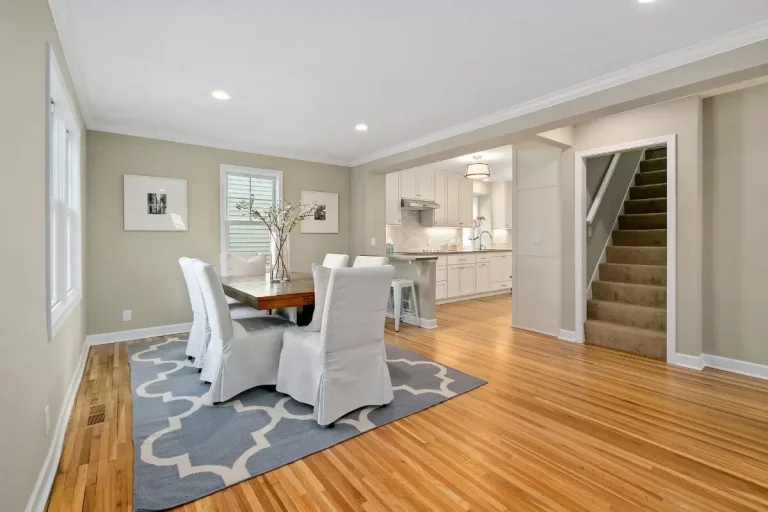 Modern dining room with hardwood floors, white chairs, blue area rug, and staircase leading to upper level