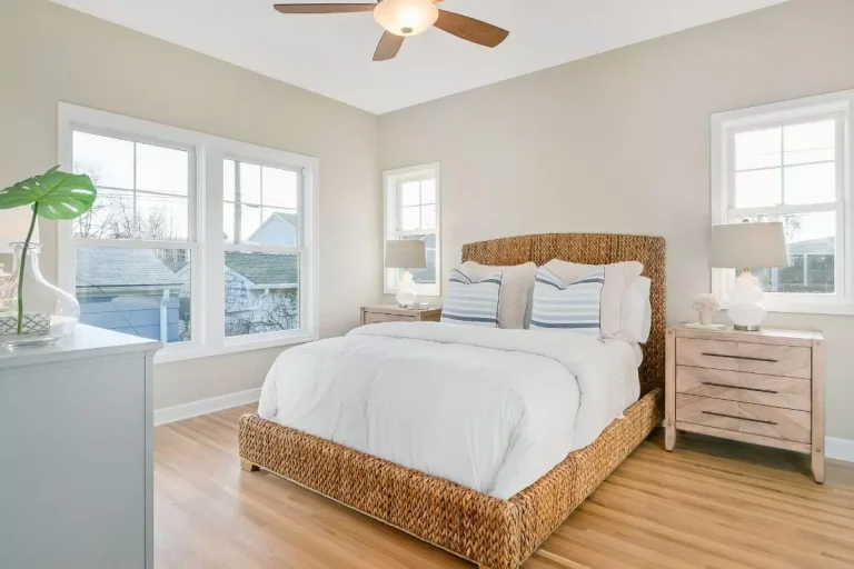 Bright, airy bedroom with wooden bed frame, white bedding, windows overlooking water, hardwood floors, and coastal decor