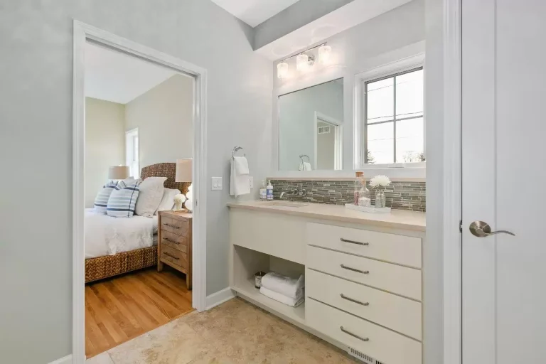 Modern bathroom with white vanity, large mirror, and open door to bedroom with hardwood floors