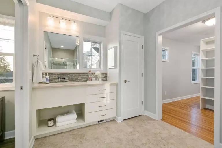Modern bathroom with white vanity, large mirror, sink, and wooden flooring leading to closet area