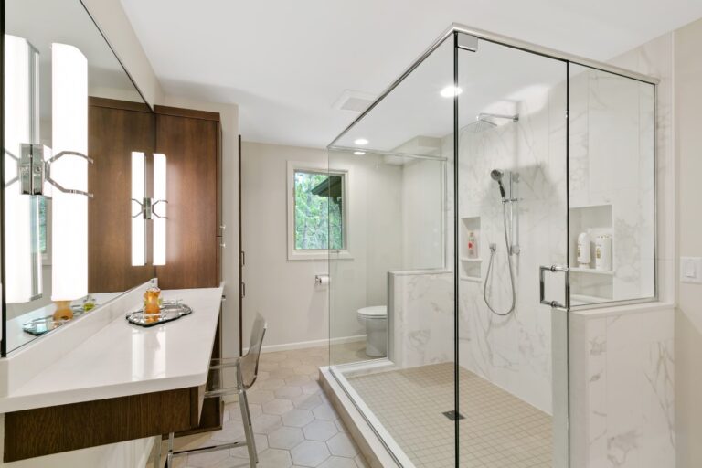 Modern luxury bathroom with large glass shower enclosure, white marble tiles, and wooden vanity with window