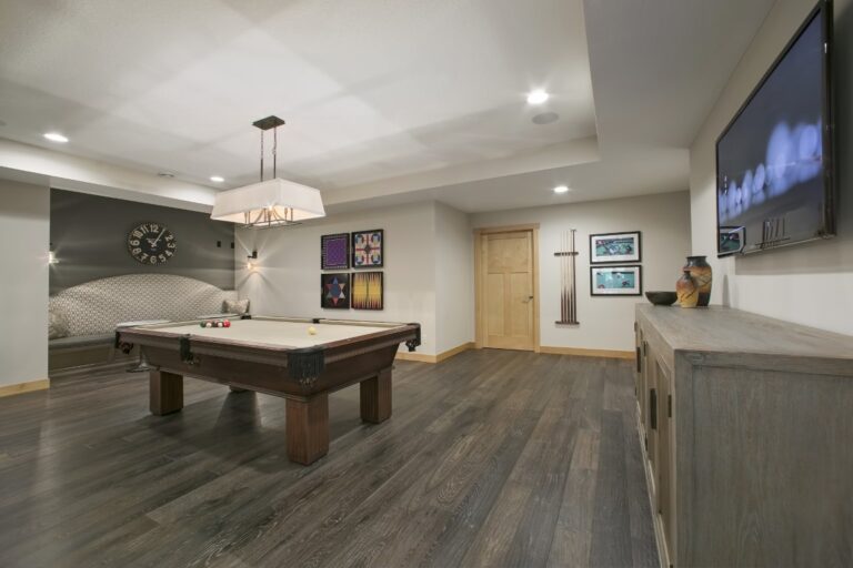 Modern basement recreation room with billiards table, bar seating, dark wood flooring, and mounted television