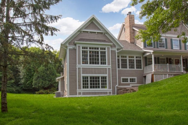 Two-story gray house with white trim and brick chimney on manicured green lawn surrounded by trees