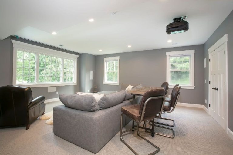 Modern home theater room with gray sectional sofa, leather armchair, projector, and large windows