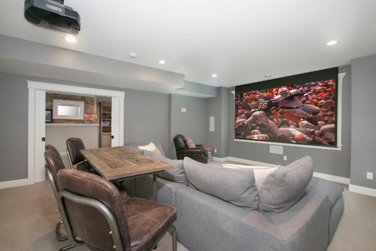 Modern home theater room with gray seating, wooden table, and wall-mounted screen displaying action scene