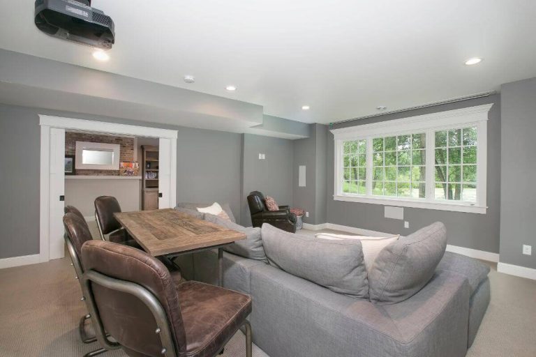 Modern home theater room with gray sectional sofa, leather armchairs, wooden coffee table, and large windows
