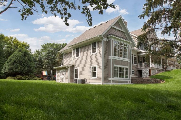 Two-story gray house with white trim on a large green lawn surrounded by mature trees on a sunny day