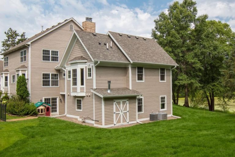 Two-story beige house with attached garage on green lawn surrounded by trees