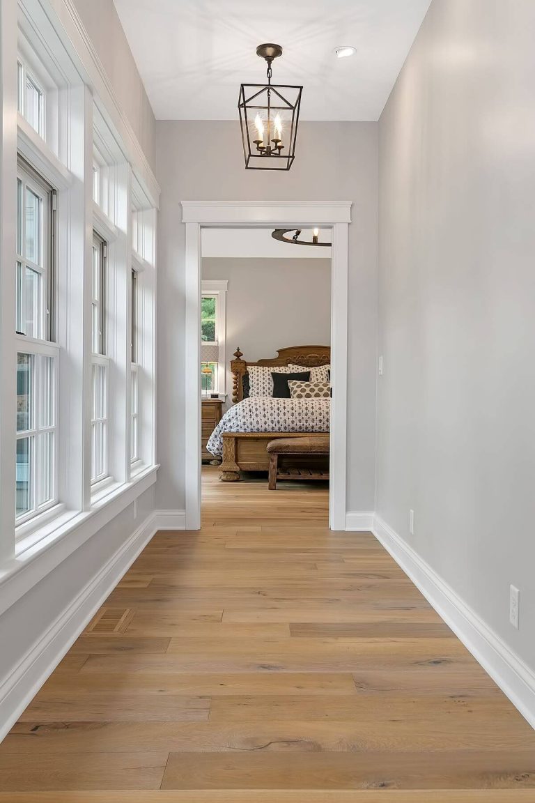 Bright hallway with white walls, hardwood flooring, windows on left, and view into bedroom with bed at end
