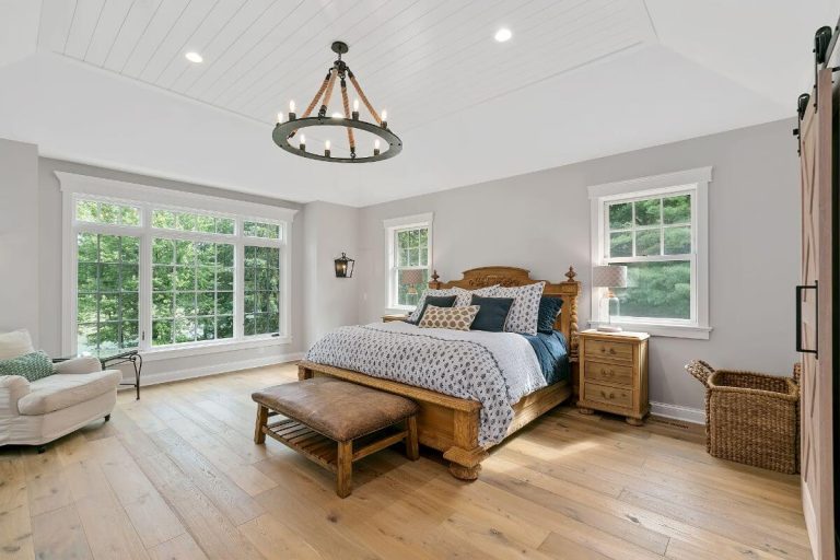 Bright master bedroom with wood flooring, blue bed, wooden bench, wicker baskets, and large windows overlooking greenery