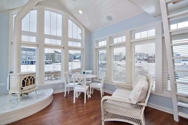 Bright sunroom with arched windows, wooden flooring, white wicker chair, small table, and water views