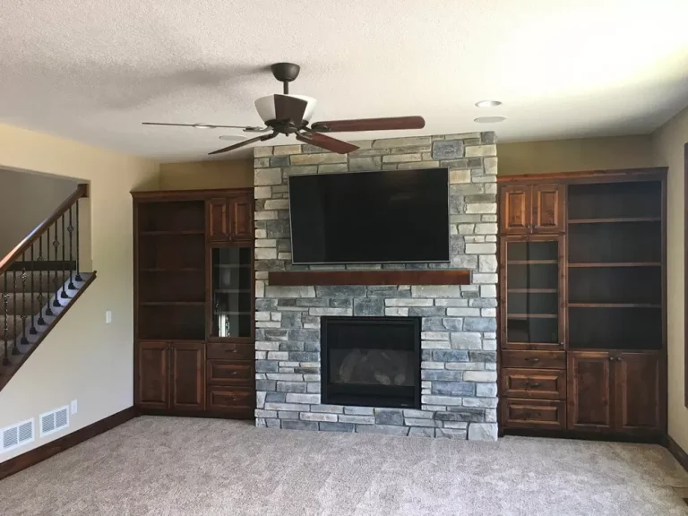 Modern living room with stone fireplace, flat screen TV, ceiling fan, wooden built-in cabinets, and staircase