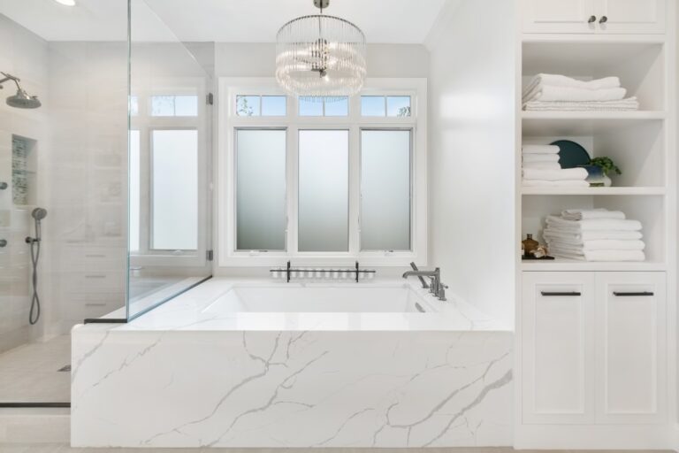 Luxurious white marble bathroom with soaking tub, large window, ceiling light fixture, and built-in white storage shelving