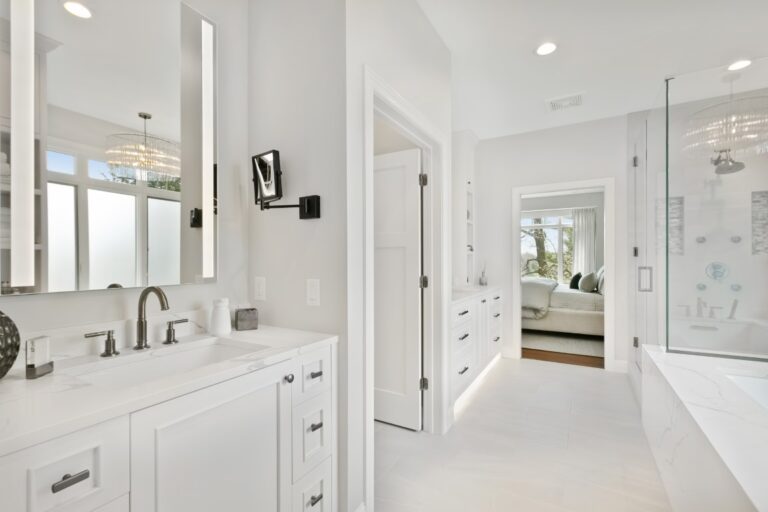 Modern white bathroom with dual vanities, marble countertops, glass shower enclosure, and neutral tile flooring