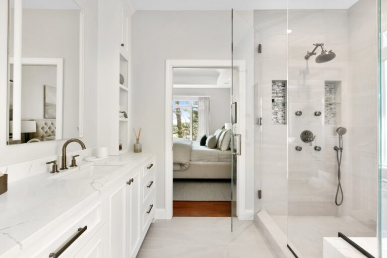 Modern luxury bathroom with white cabinetry, double vanity, walk-in shower, and view into bedroom beyond