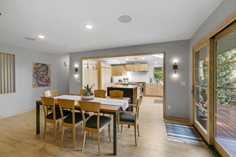 Modern open-concept dining and kitchen area with wooden table, mid-century chairs, gray walls, and glass doors leading to