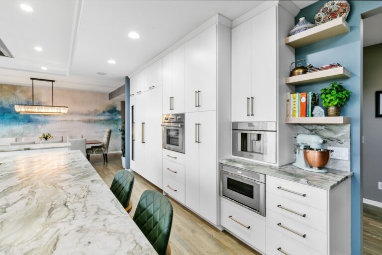 Modern white kitchen with marble countertops, stainless steel appliances, blue open shelving, and dining area with green