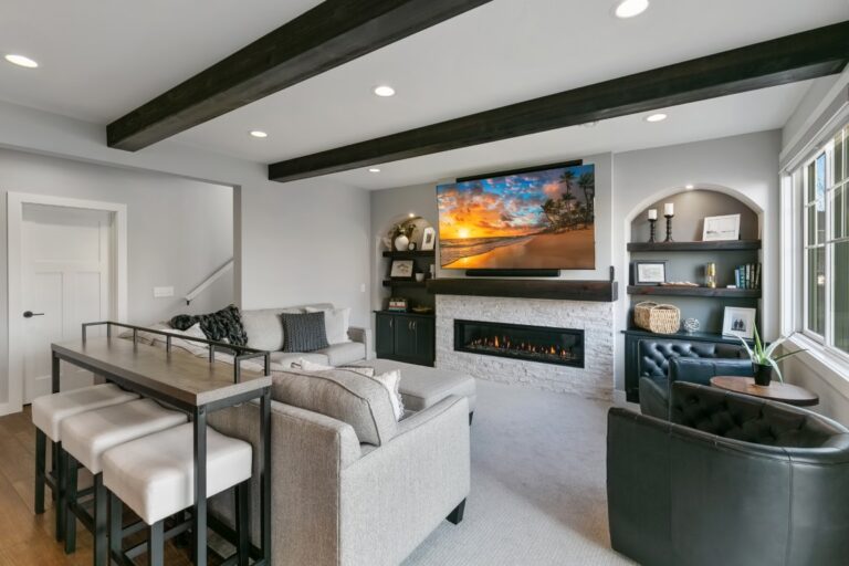Modern living room with fireplace, built-in shelving, white furniture, dark wood ceiling beams, and landscape artwork