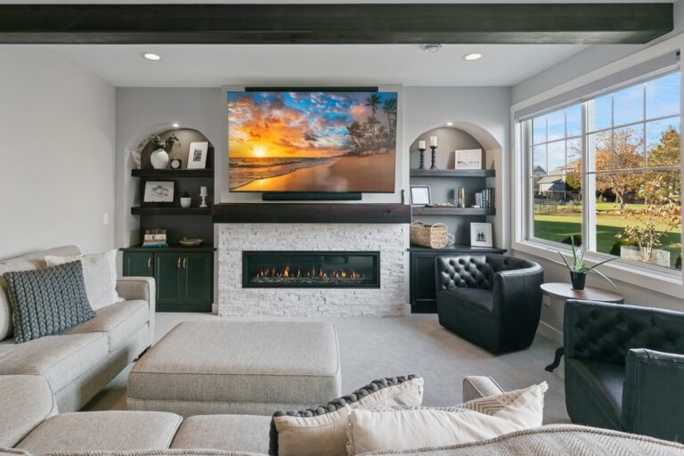 Modern living room with fireplace, TV, black leather seating, shelving, and large windows overlooking landscaped yard