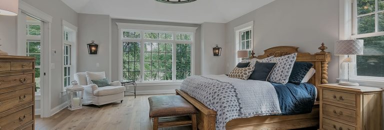Bright bedroom with wooden furniture, large windows, seating area, and neutral bedding